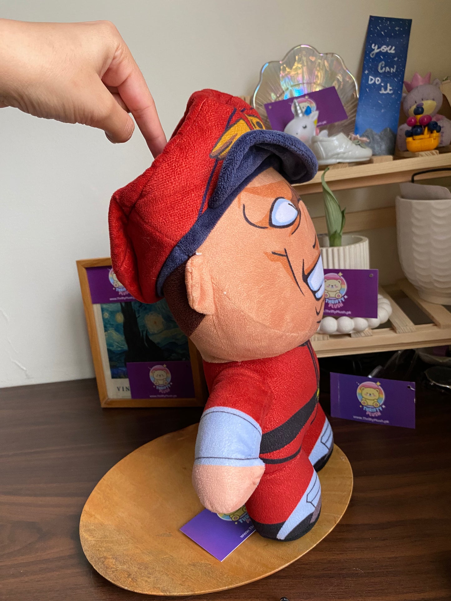 A plush toy of E. Honda from Street Fighter, dressed in his signature red sumo outfit with a blue and red cap, is displayed on a wooden surface. The character's face is depicted with a wide, toothy grin and prominent facial features. In the background, a framed picture and a shelf with various decorative items, including a "you can do it" sign, are visible