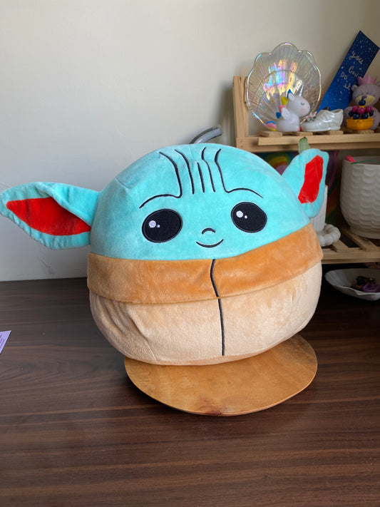 Plush toy resembling Baby Yoda from 'The Mandalorian' on a wooden surface with a blurred background.