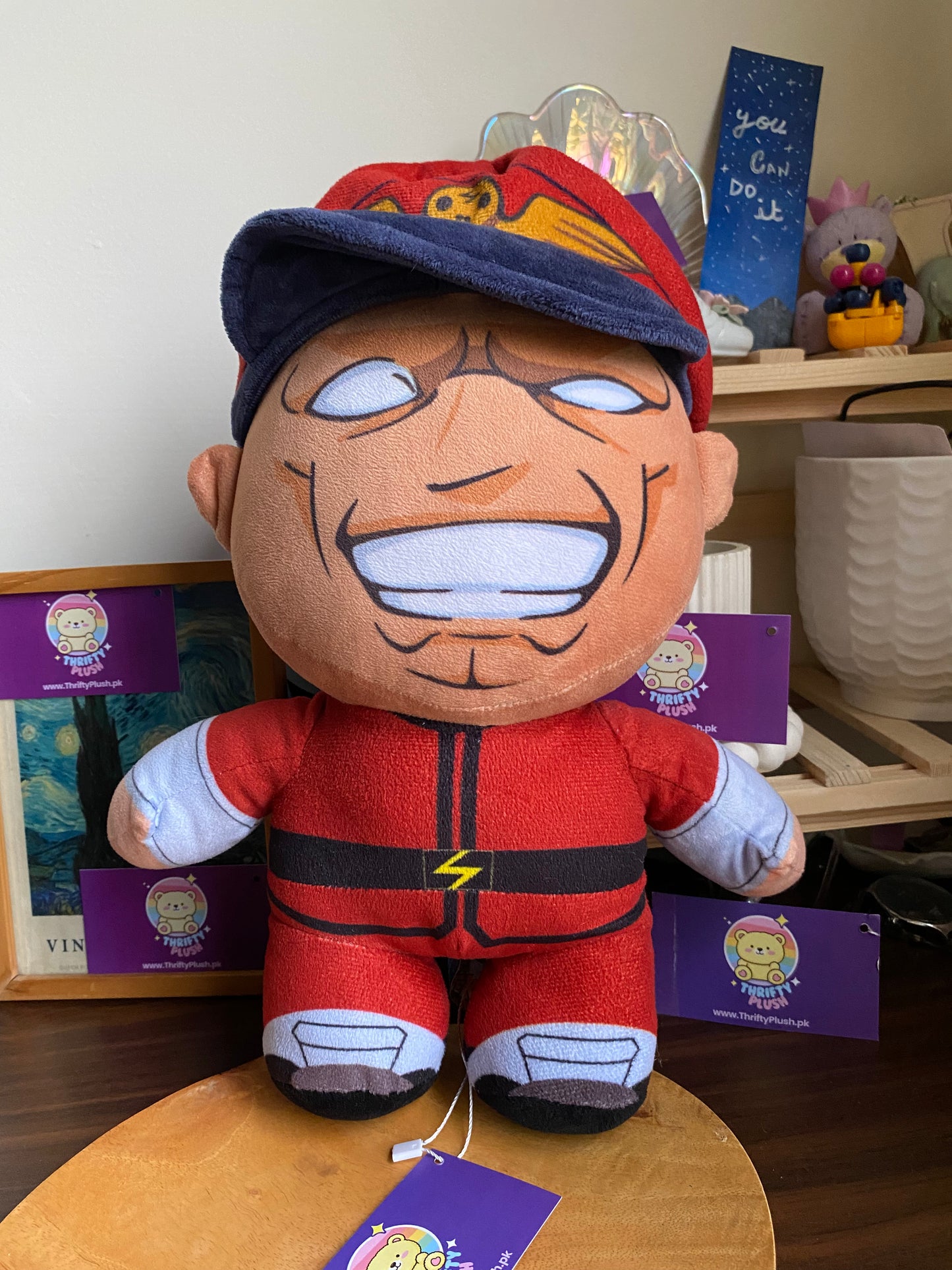 A plush toy of E. Honda from Street Fighter, dressed in his signature red sumo outfit with a blue and red cap, is displayed on a wooden surface. The character's face is depicted with a wide, toothy grin and prominent facial features. In the background, a framed picture and a shelf with various decorative items, including a "you can do it" sign, are visible