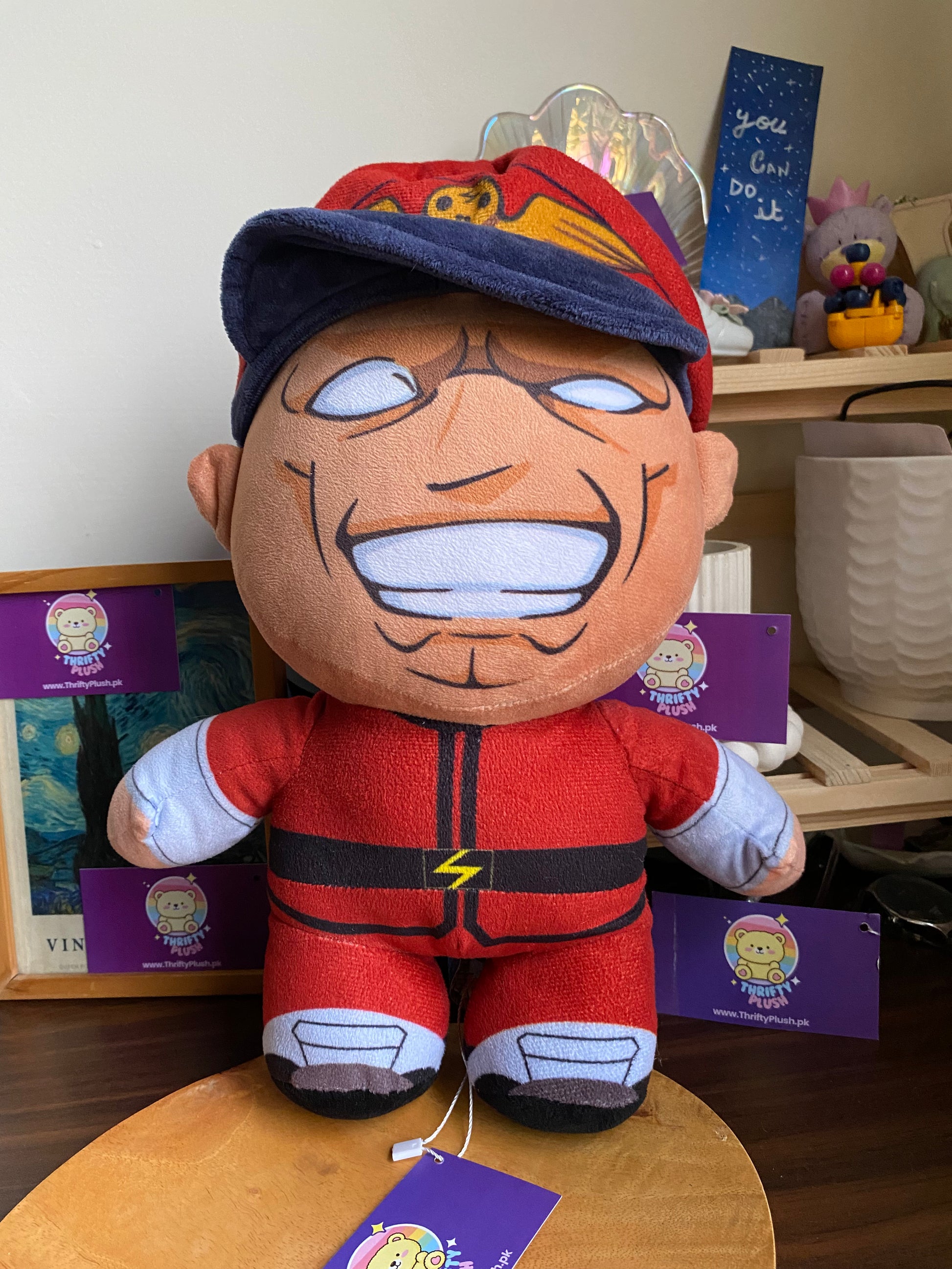 A plush toy of E. Honda from Street Fighter, dressed in his signature red sumo outfit with a blue and red cap, is displayed on a wooden surface. The character's face is depicted with a wide, toothy grin and prominent facial features. In the background, a framed picture and a shelf with various decorative items, including a "you can do it" sign, are visible