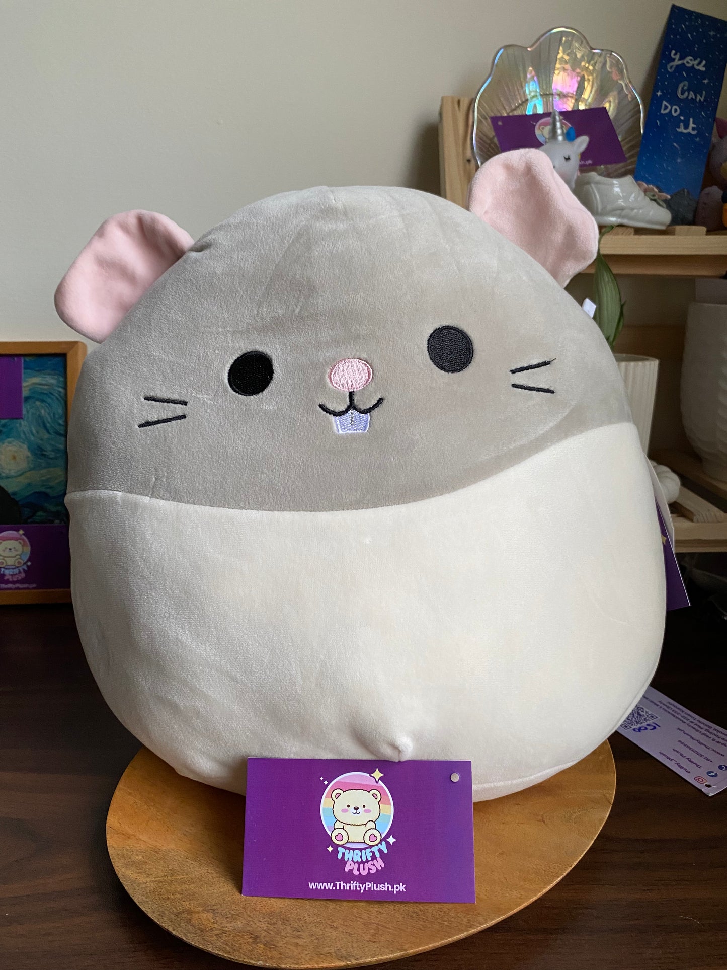 large, round, grey and white Squishmallows plush toy named Rusty, designed to resemble a rat or mouse, sitting on a wooden surface
