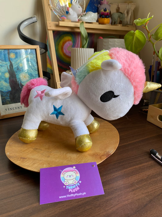 Unicorn plush toy with rainbow hair on a wooden surface, next to a purple card with branding.