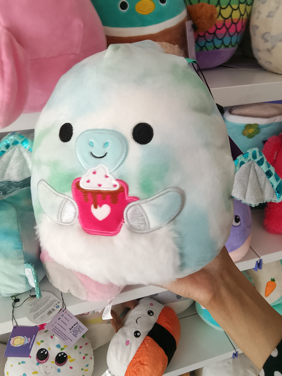 Squishmallows Shantira the Pterodactyl plush toy blue and white tie-dye design and holding a hot chocolate cup.