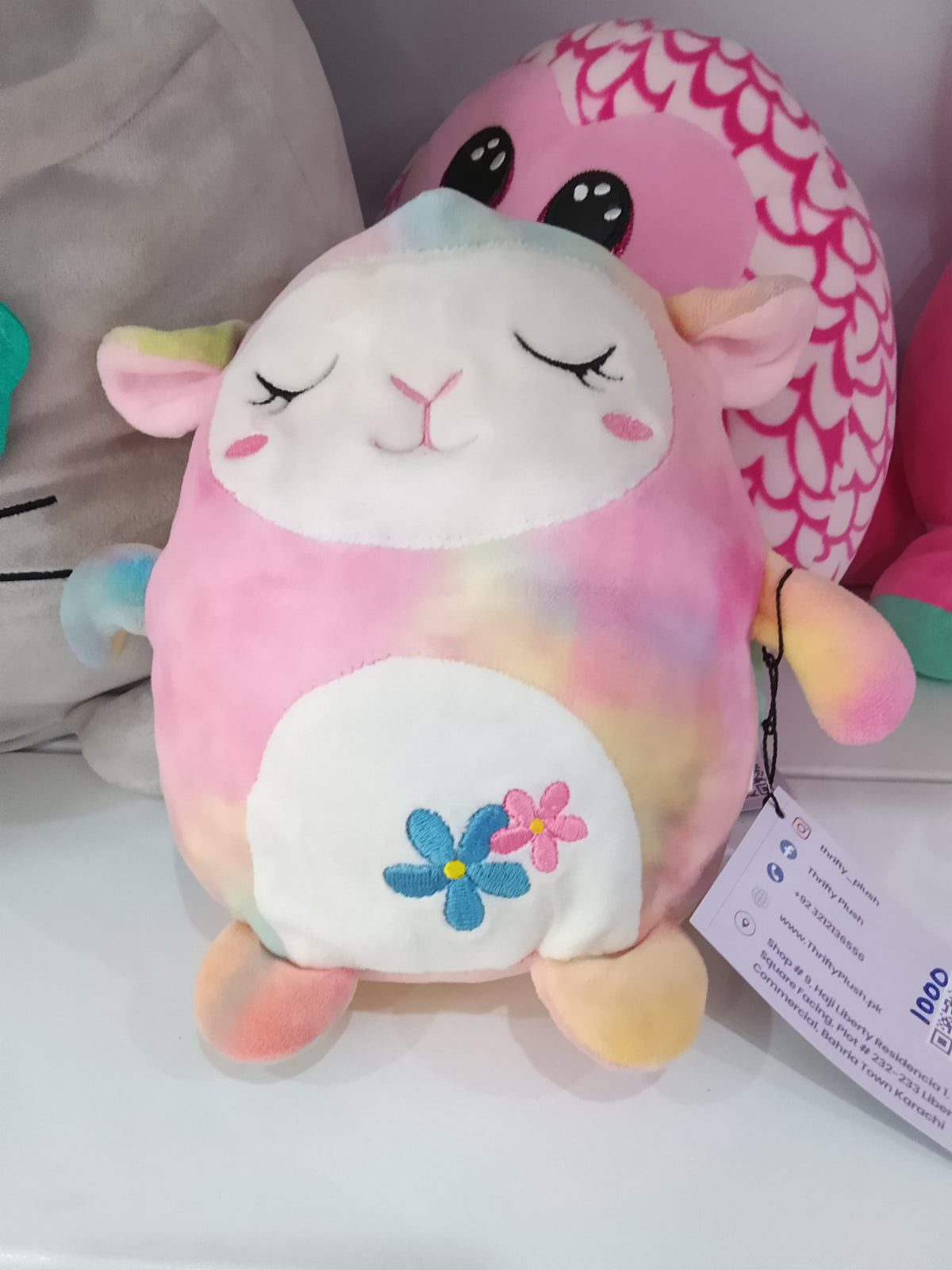 Squishmallows Lana the rainbow lamb plushie, featuring a colorful tie-dye design with embroidered flowers.