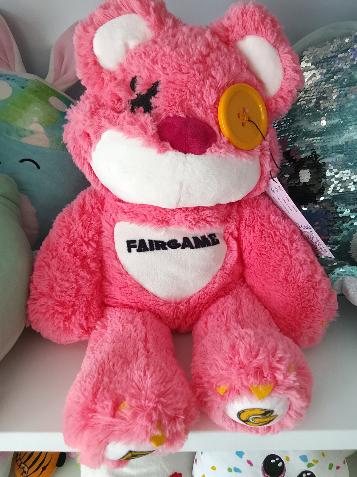 Fairgame brand pink teddy bear plush toy with a yellow button eye and a stitched "X" eye.